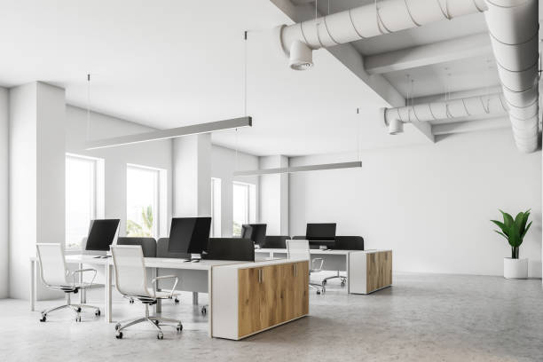 Corner of an open space office with white walls, a concrete floor and rows of computer tables. 3d rendering mock up